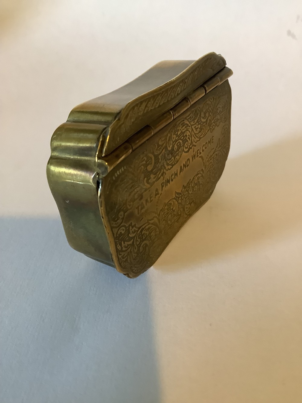 An antique hinged brass snuff box featuring ornate foliate scrollwork engraving and the central inscription reading TAKE A PINCH AND WELCOME. View V