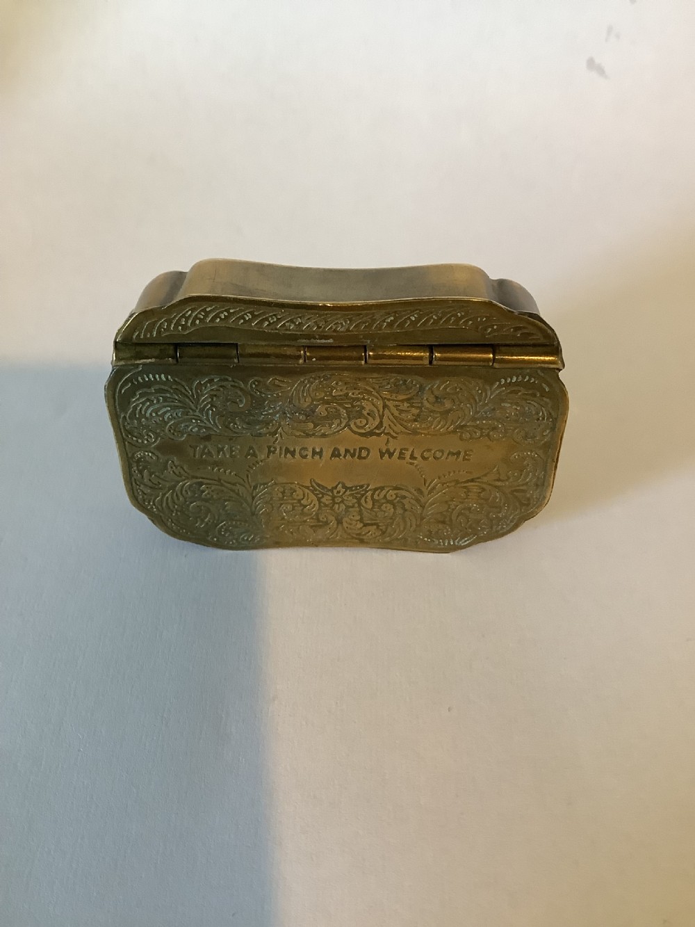 An antique hinged brass snuff box featuring ornate foliate scrollwork engraving and the central inscription reading TAKE A PINCH AND WELCOME. View VI