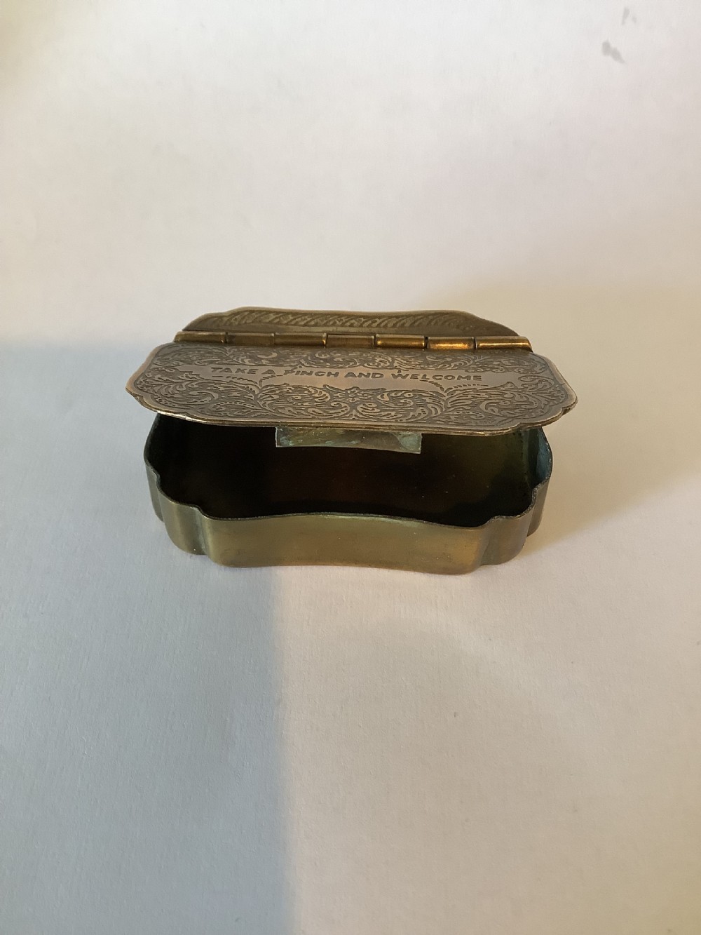 An antique hinged brass snuff box featuring ornate foliate scrollwork engraving and the central inscription reading TAKE A PINCH AND WELCOME. View VII