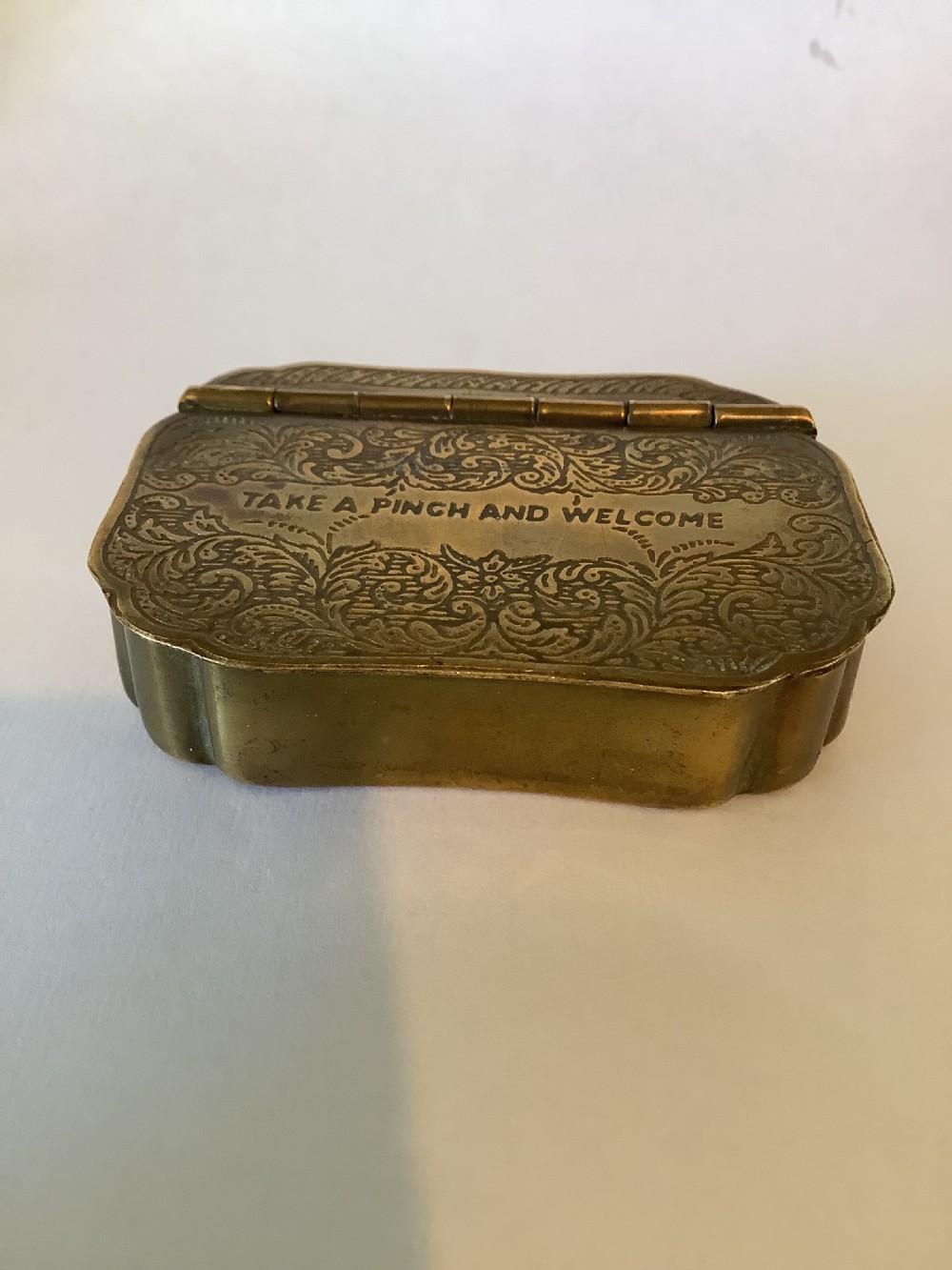 An antique hinged brass snuff box featuring ornate foliate scrollwork engraving and the central inscription reading TAKE A PINCH AND WELCOME. View VIII