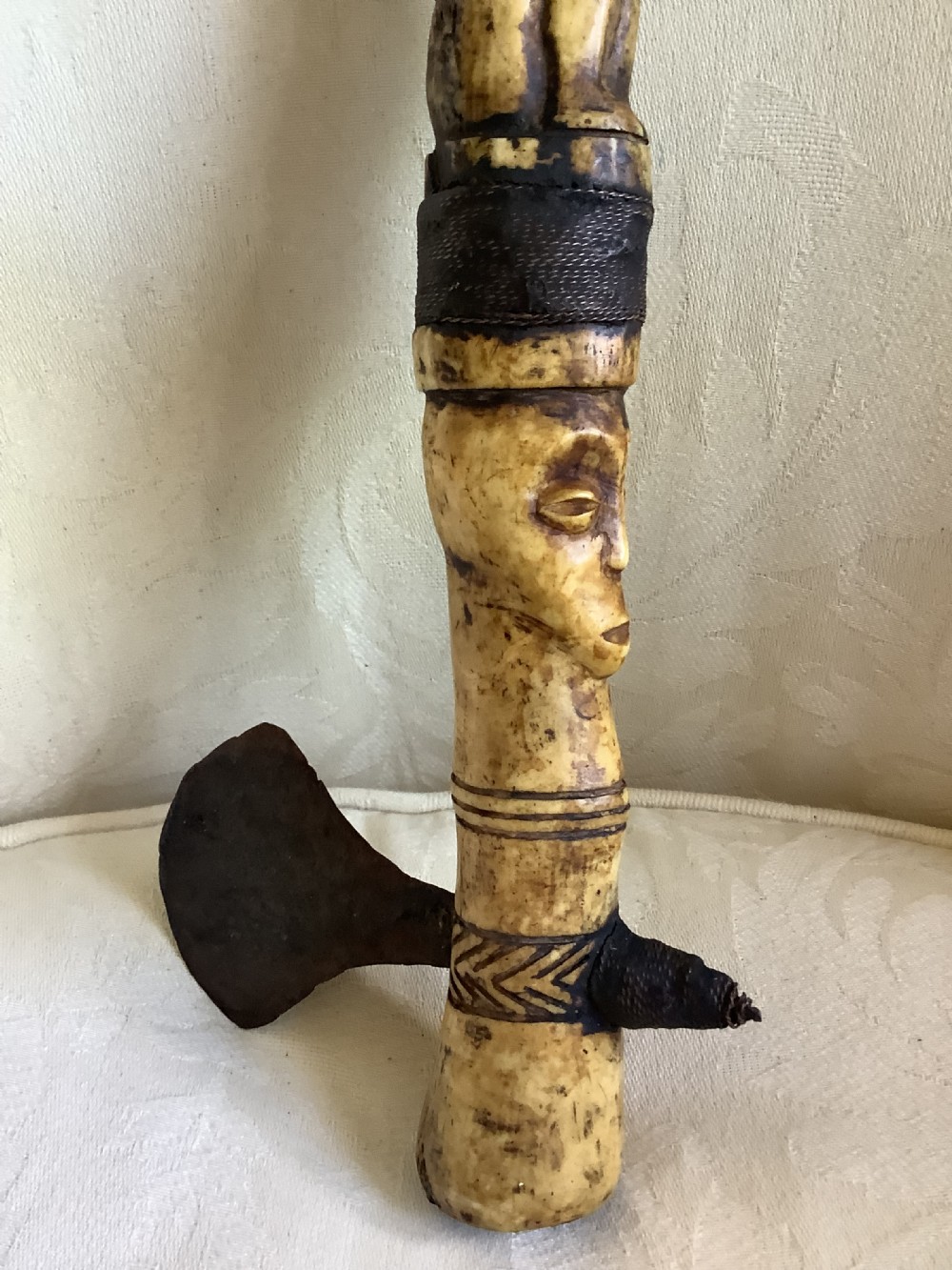 A carved bone ceremonial axe featuring stacked figurative human carvings along the shaft and a small dark iron blade fixed near the base. View VII