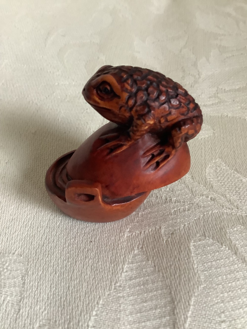 A dark brown carved wood netsuke shaped as a textured toad resting on a curved base. View V