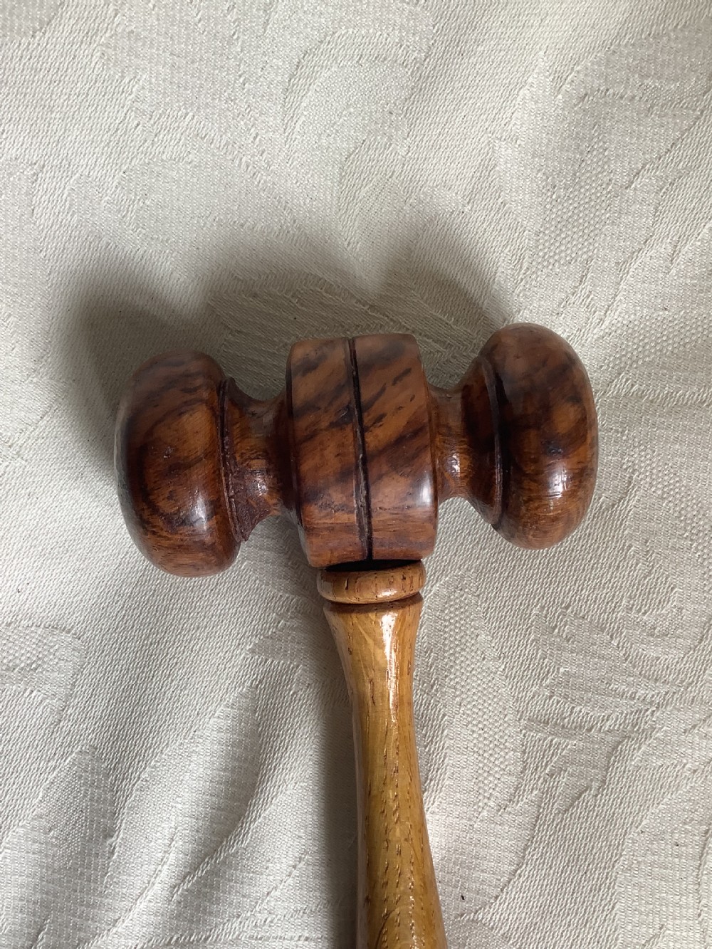 A hand-turned brown wooden gavel featuring a double-ended striking head and an incised handle resting on a textured light fabric background. View II