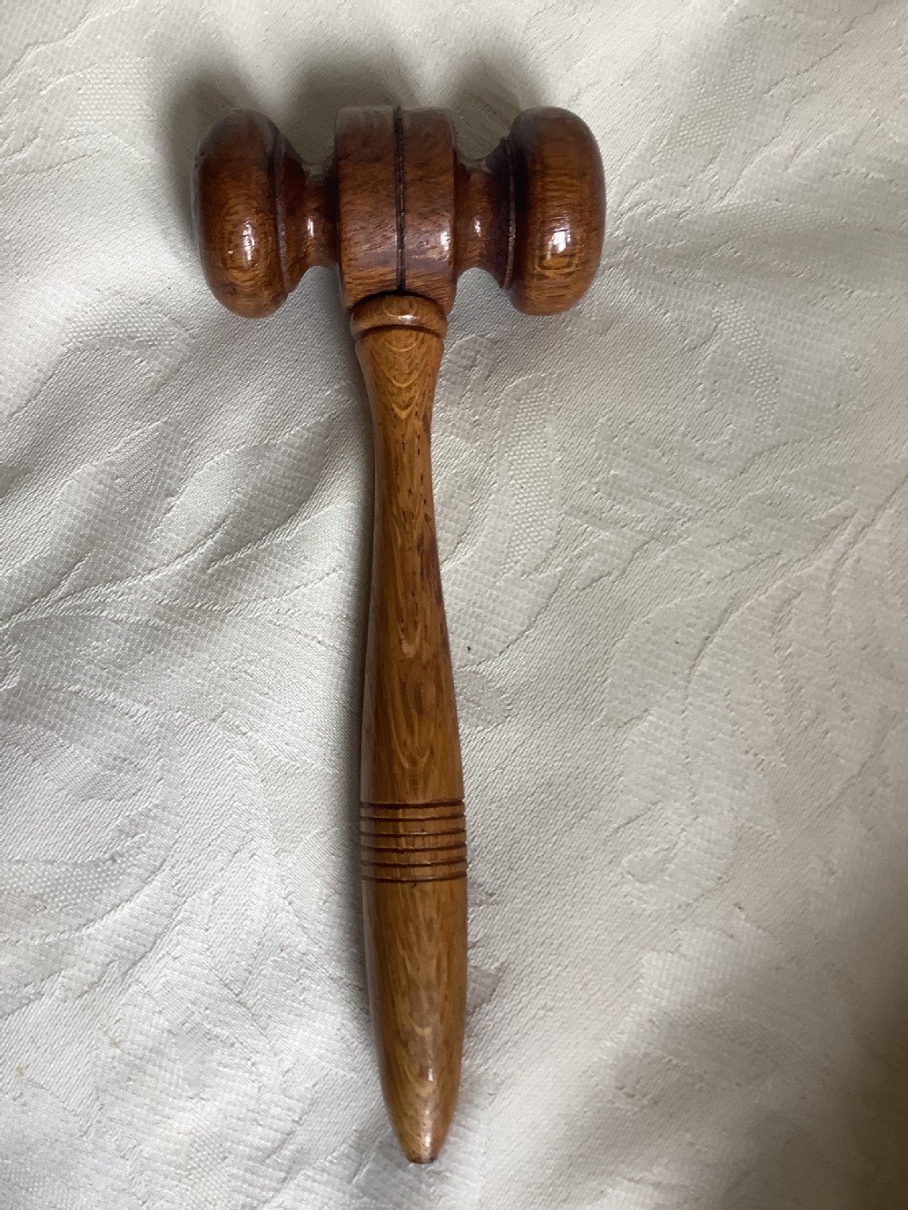 A hand-turned brown wooden gavel featuring a double-ended striking head and an incised handle resting on a textured light fabric background. View III