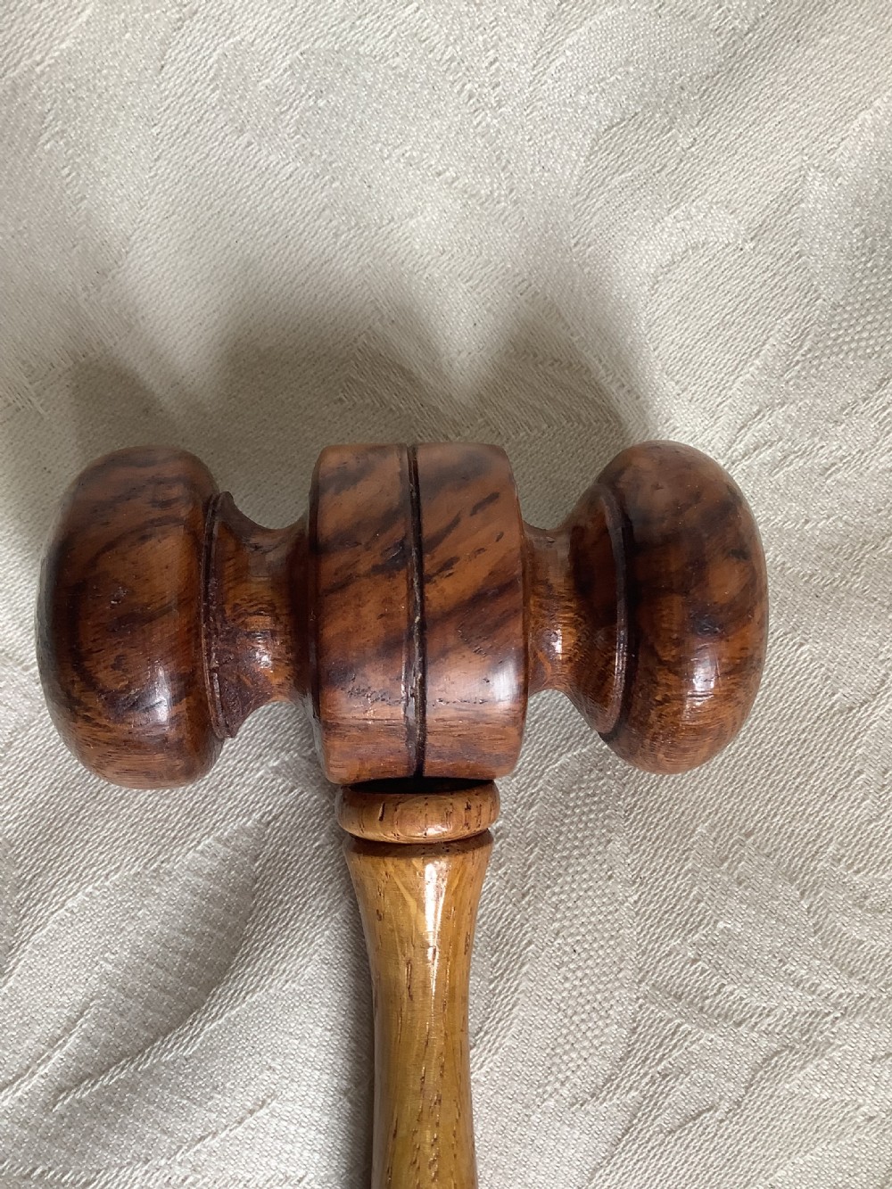A hand-turned brown wooden gavel featuring a double-ended striking head and an incised handle resting on a textured light fabric background. View IV
