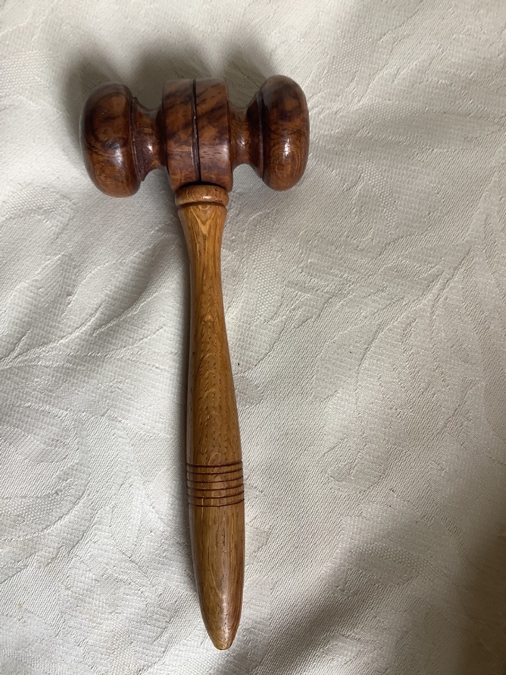 A hand-turned brown wooden gavel featuring a double-ended striking head and an incised handle resting on a textured light fabric background. View V