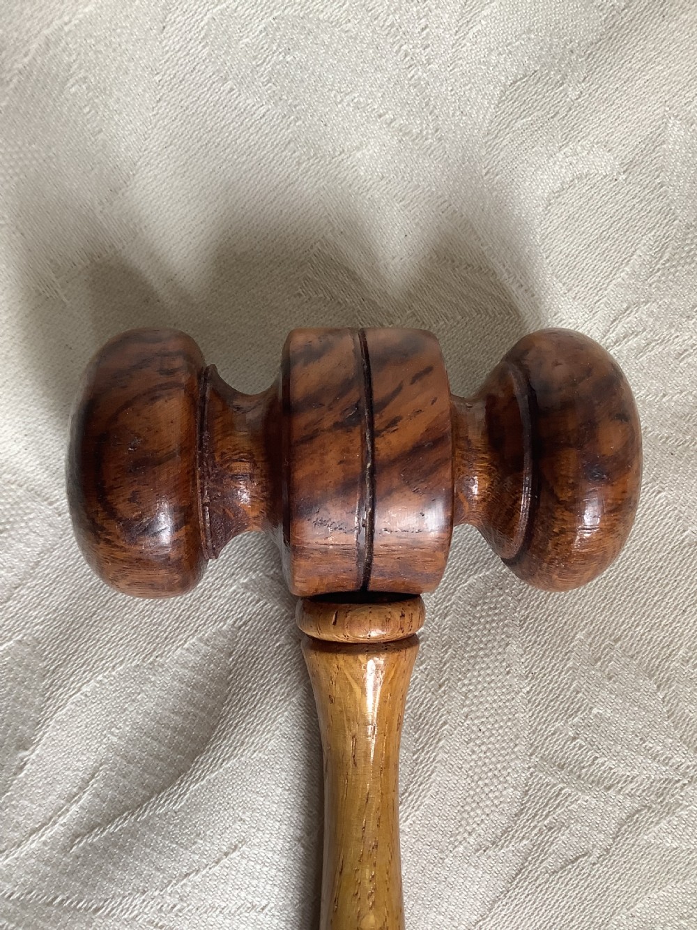 A hand-turned brown wooden gavel featuring a double-ended striking head and an incised handle resting on a textured light fabric background. View VI