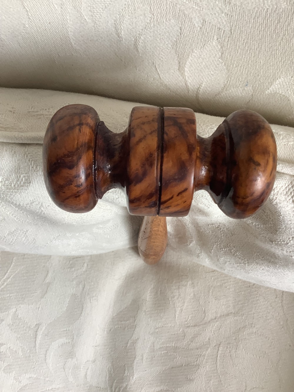 A hand-turned brown wooden gavel featuring a double-ended striking head and an incised handle resting on a textured light fabric background. View VII