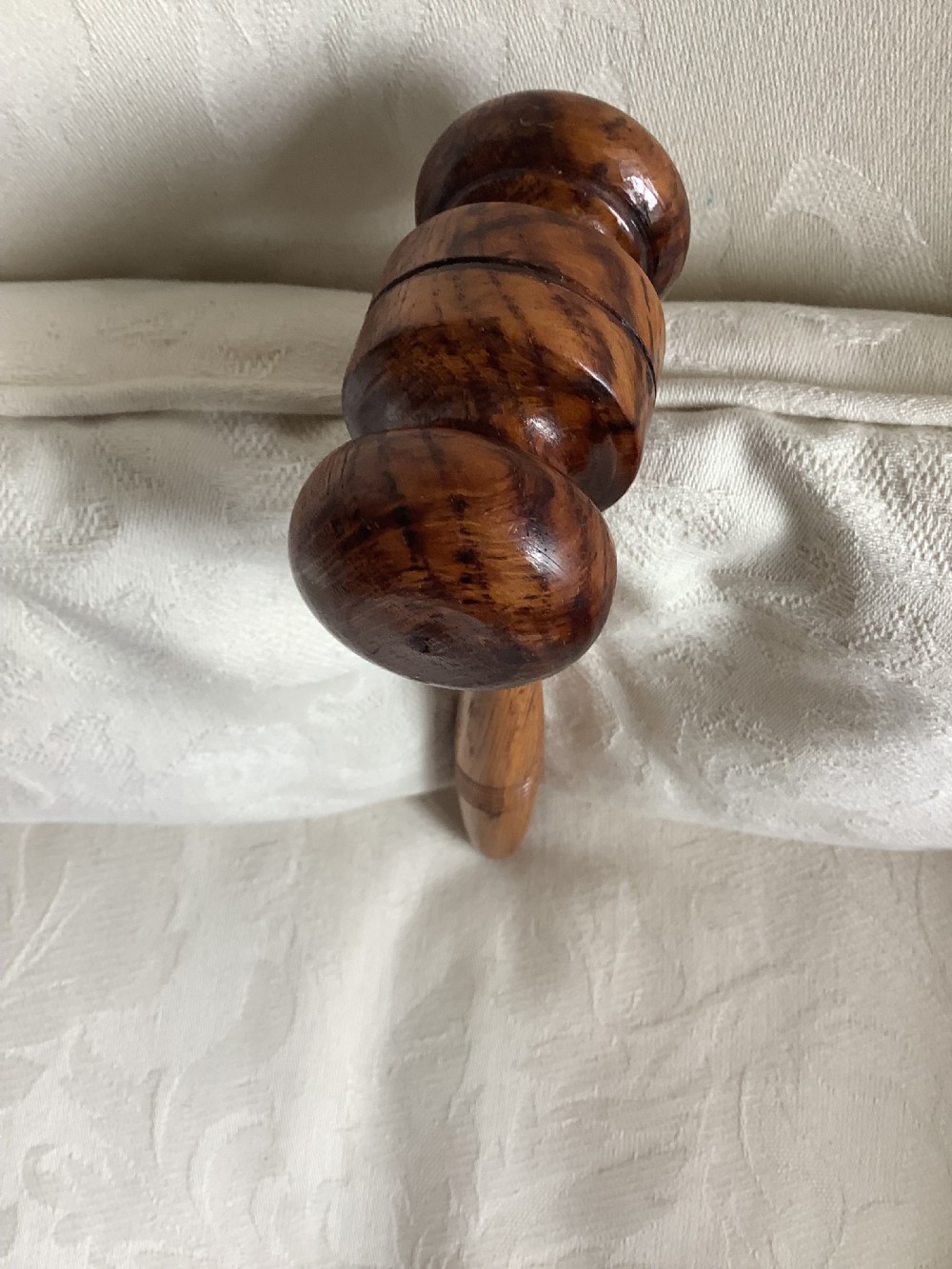 A hand-turned brown wooden gavel featuring a double-ended striking head and an incised handle resting on a textured light fabric background. View VIII