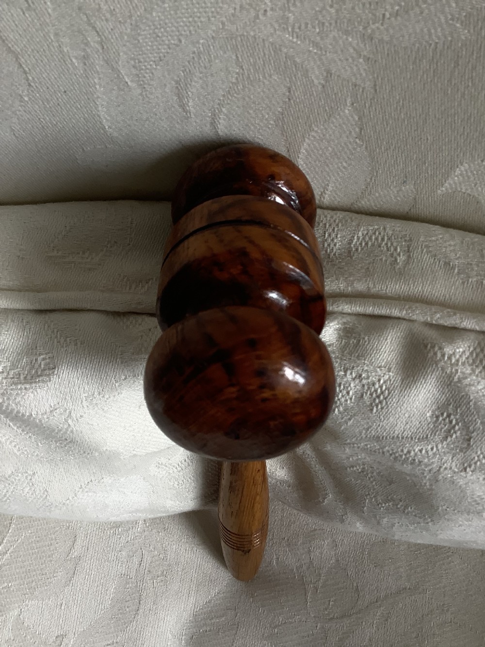 A hand-turned brown wooden gavel featuring a double-ended striking head and an incised handle resting on a textured light fabric background. View IX