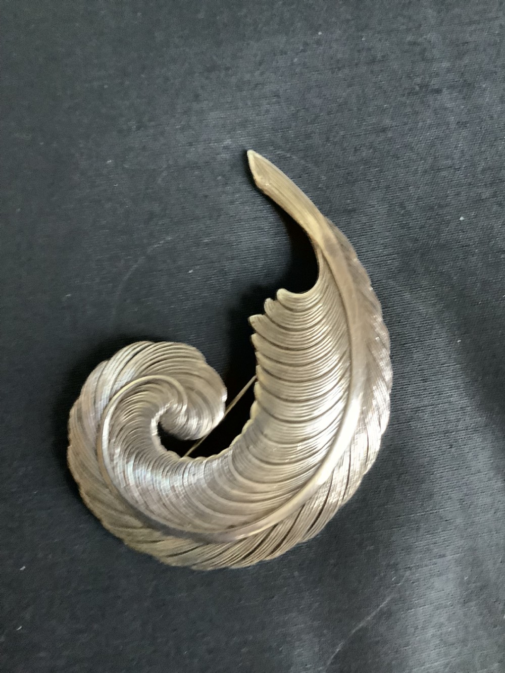 A curved white metal brooch designed in the shape of a textured feather plume with a pin fastening on the reverse. View II