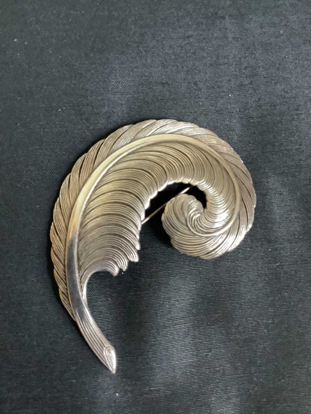 A curved white metal brooch designed in the shape of a textured feather plume with a pin fastening on the reverse. View III