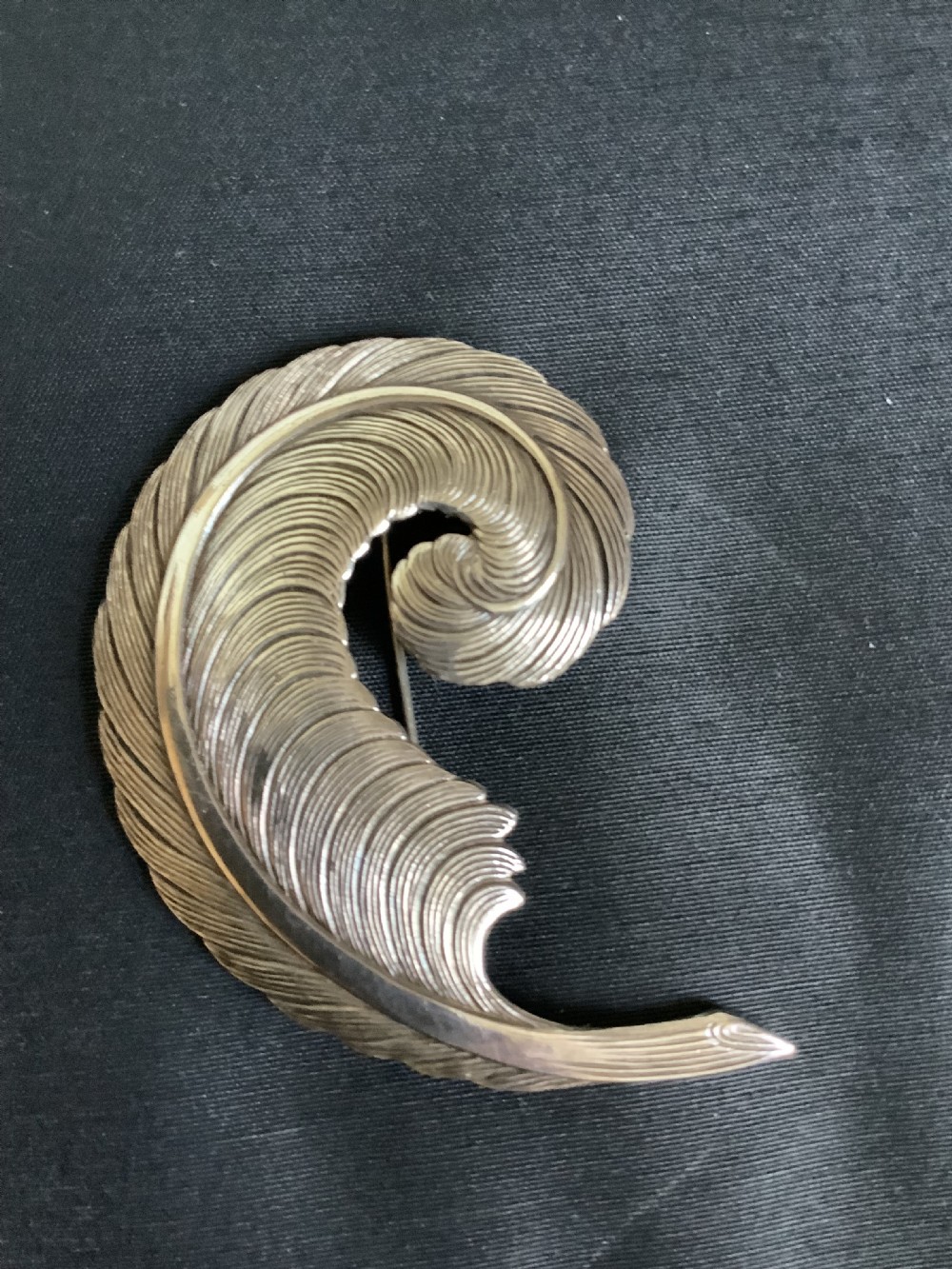 A curved white metal brooch designed in the shape of a textured feather plume with a pin fastening on the reverse. View VI