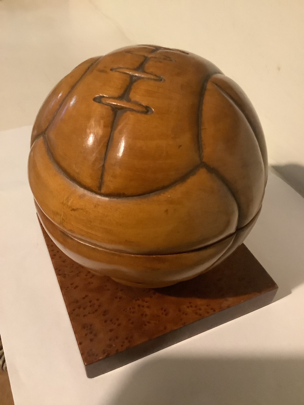 A carved hardwood lidded box shaped like a vintage laced football, mounted on a square burr wood base.