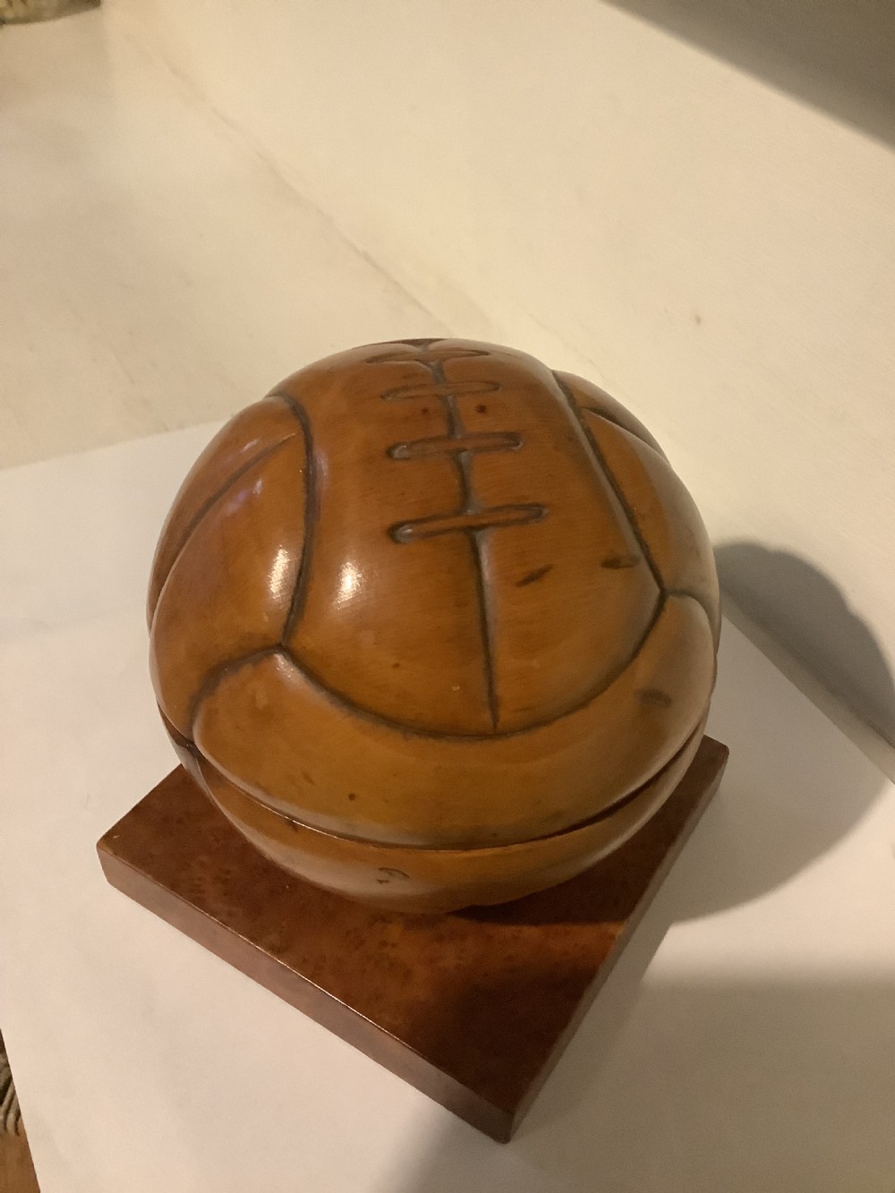 A carved hardwood lidded box shaped like a vintage laced football, mounted on a square burr wood base. View V