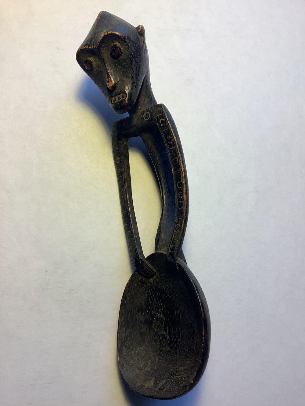 A dark carved wooden spoon featuring a handle sculpted into a stylized human figure with incised geometric markings along the arms.
