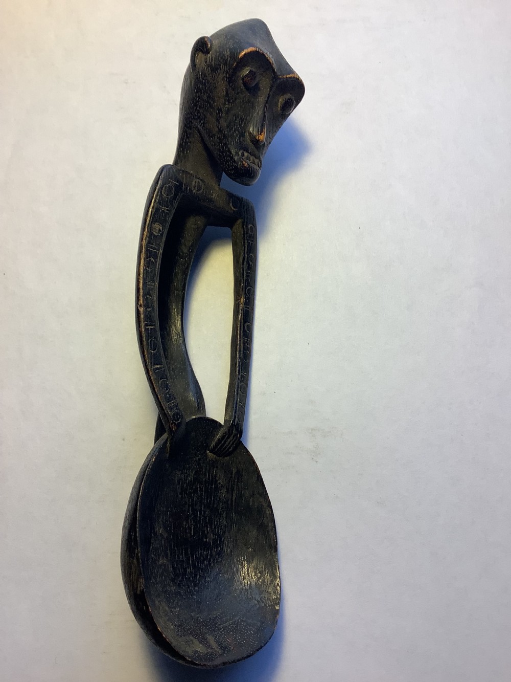 A dark carved wooden spoon featuring a handle sculpted into a stylized human figure with incised geometric markings along the arms. View II