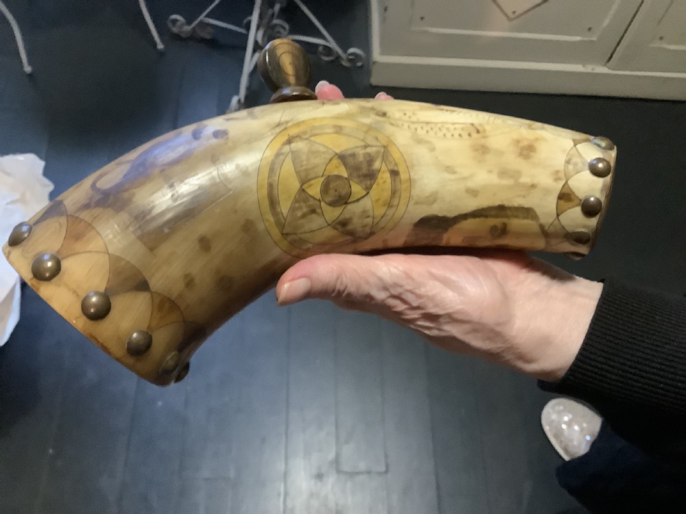 A curved antique powder horn featuring geometric scrimshaw engravings, brass studs along the borders, and a turned wooden plug.
