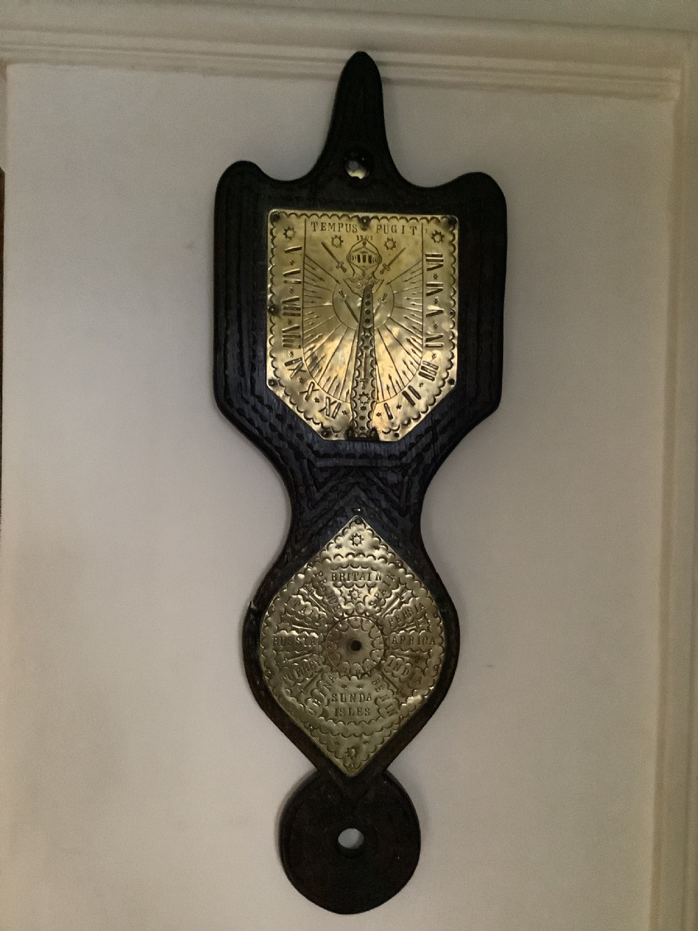 An antique brass sundial and world clock plaque mounted on a carved dark wooden backboard, featuring engraved Roman numerals and global place names.
