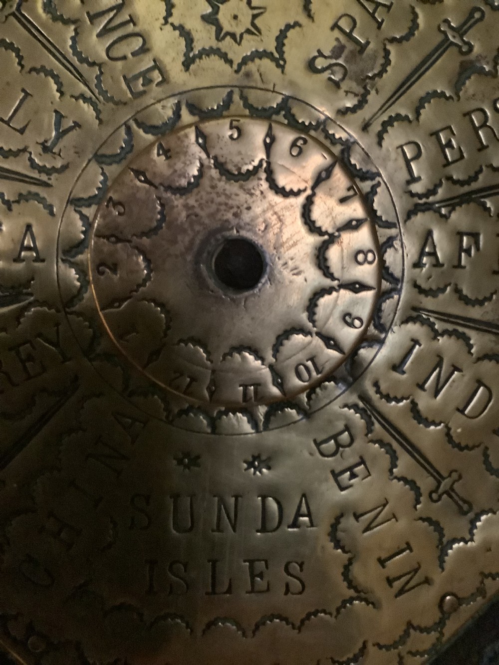 An antique brass sundial and world clock plaque mounted on a carved dark wooden backboard, featuring engraved Roman numerals and global place names. View III