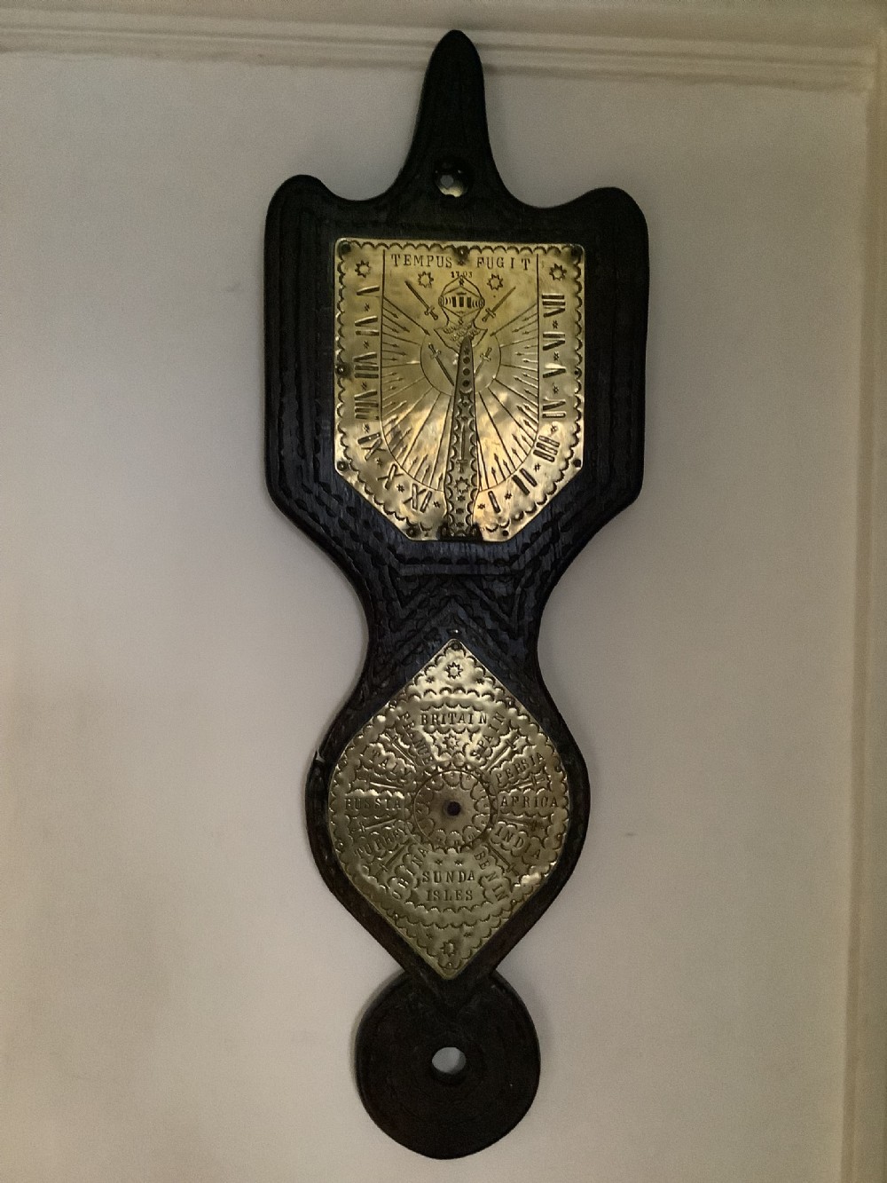 An antique brass sundial and world clock plaque mounted on a carved dark wooden backboard, featuring engraved Roman numerals and global place names. View VI