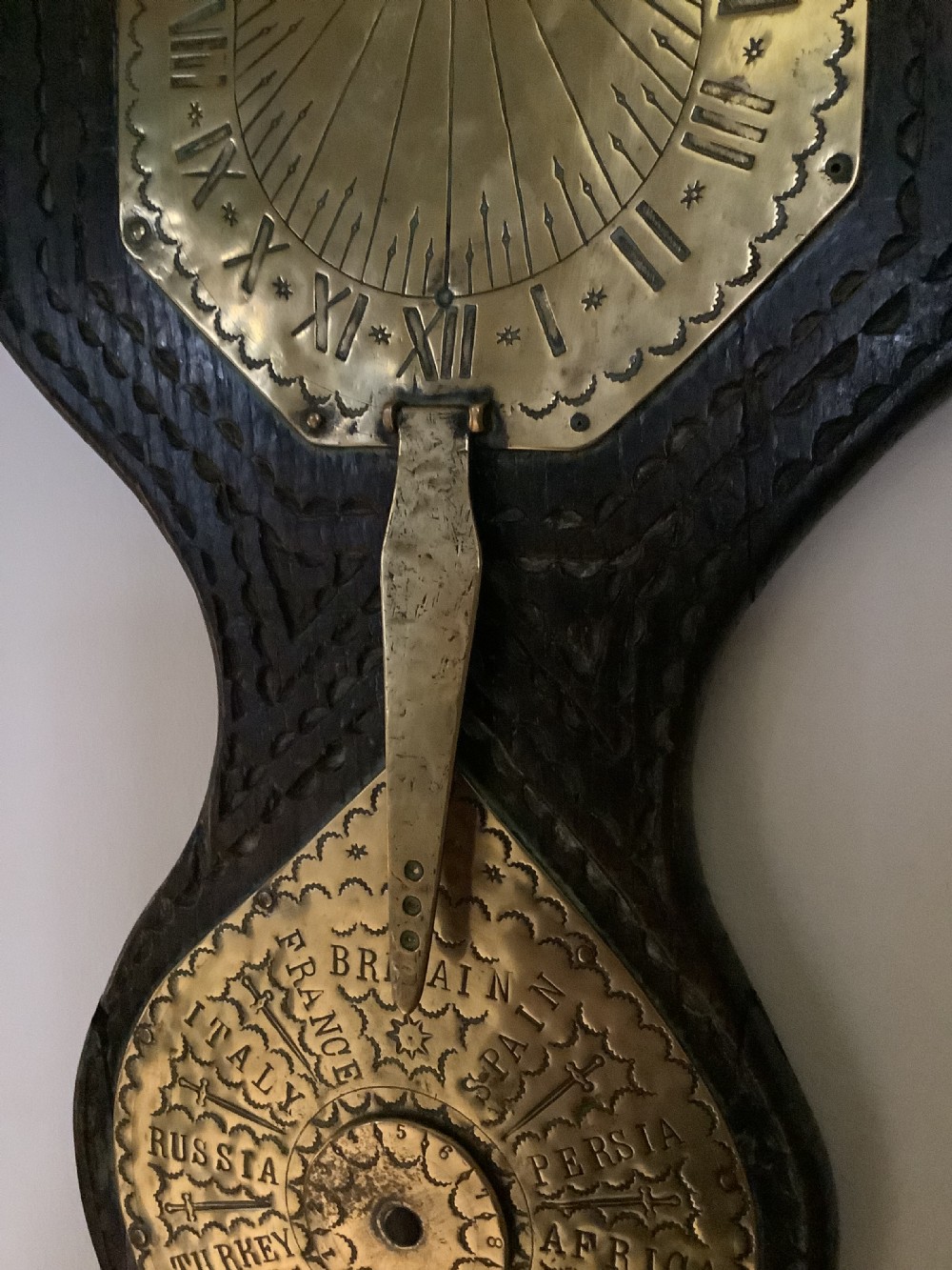 An antique brass sundial and world clock plaque mounted on a carved dark wooden backboard, featuring engraved Roman numerals and global place names. View IX