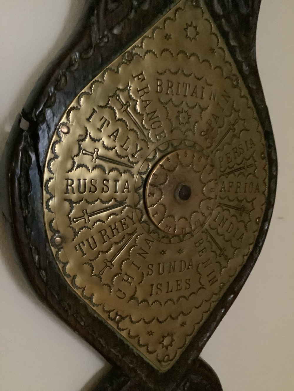 An antique brass sundial and world clock plaque mounted on a carved dark wooden backboard, featuring engraved Roman numerals and global place names. View XI