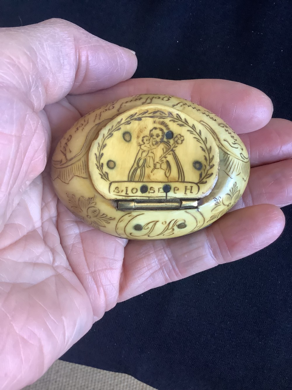 An oval antique horn pocket snuff box featuring hand-engraved depictions of Saint Joseph, floral motifs, and a central brass hinge.