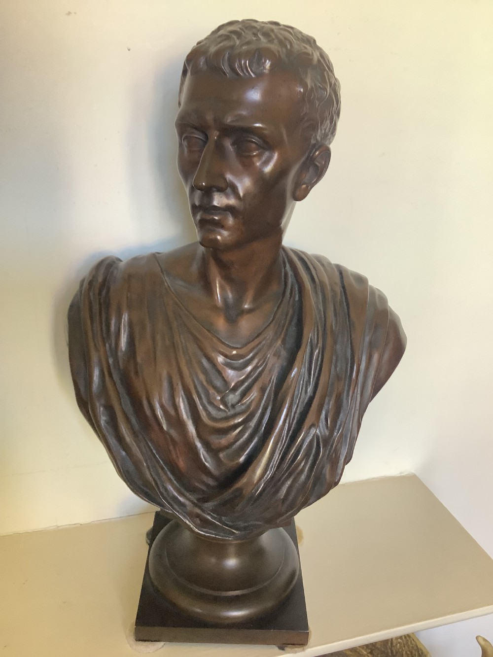 A patinated dark bronze bust depicting a classical Roman emperor with short curled hair and a draped toga, mounted on a turned circular socle and square base.