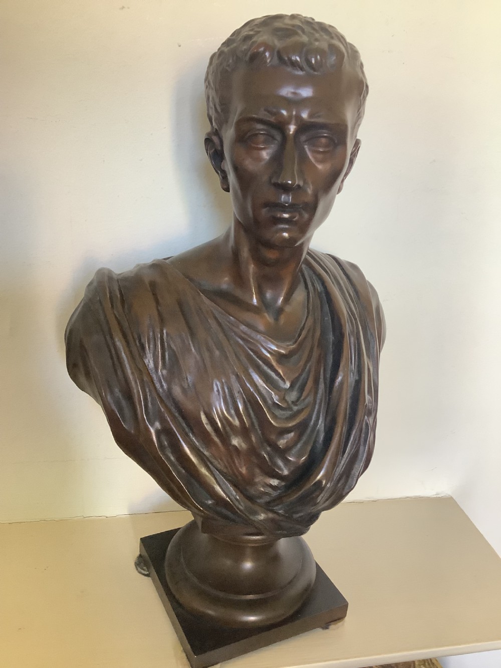 A patinated dark bronze bust depicting a classical Roman emperor with short curled hair and a draped toga, mounted on a turned circular socle and square base. View V