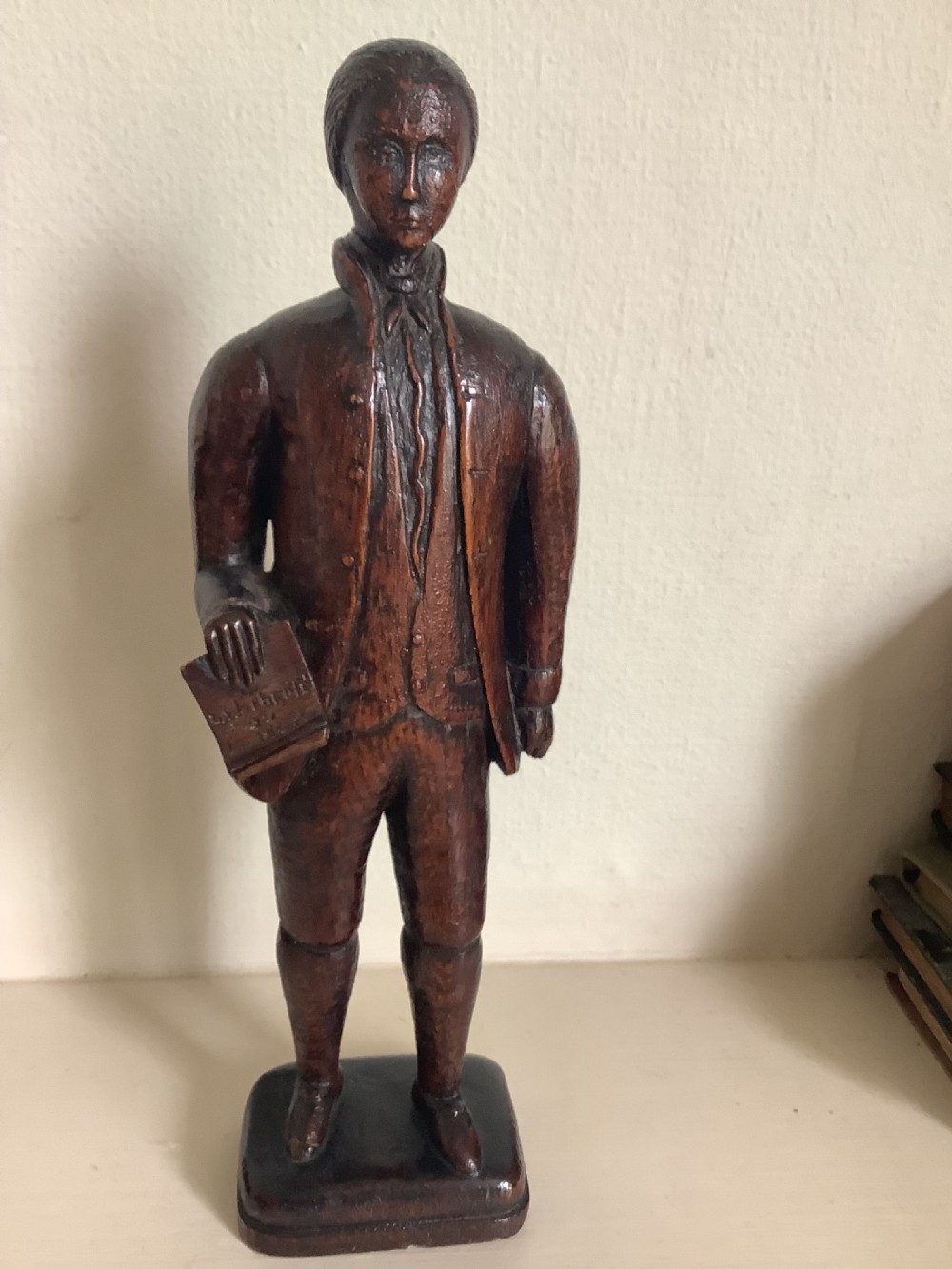 A dark hand-carved wood figure of an 18th-century man holding a book, standing on an integrated wooden base.