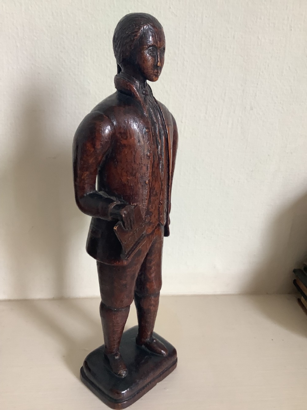 A dark hand-carved wood figure of an 18th-century man holding a book, standing on an integrated wooden base. View II