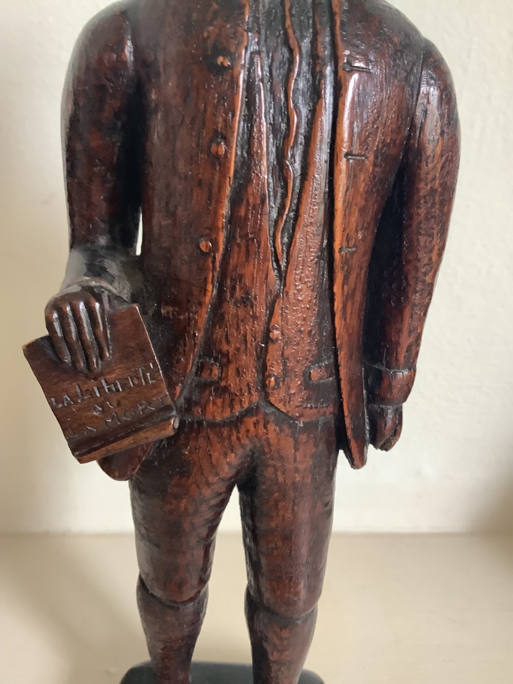 A dark hand-carved wood figure of an 18th-century man holding a book, standing on an integrated wooden base. View III
