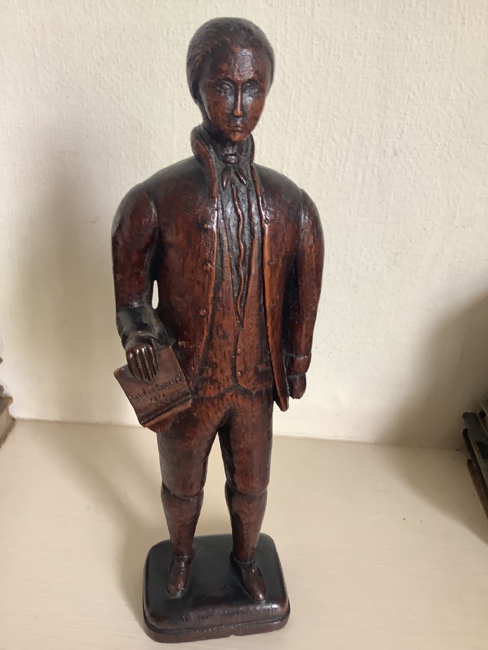 A dark hand-carved wood figure of an 18th-century man holding a book, standing on an integrated wooden base. View IV