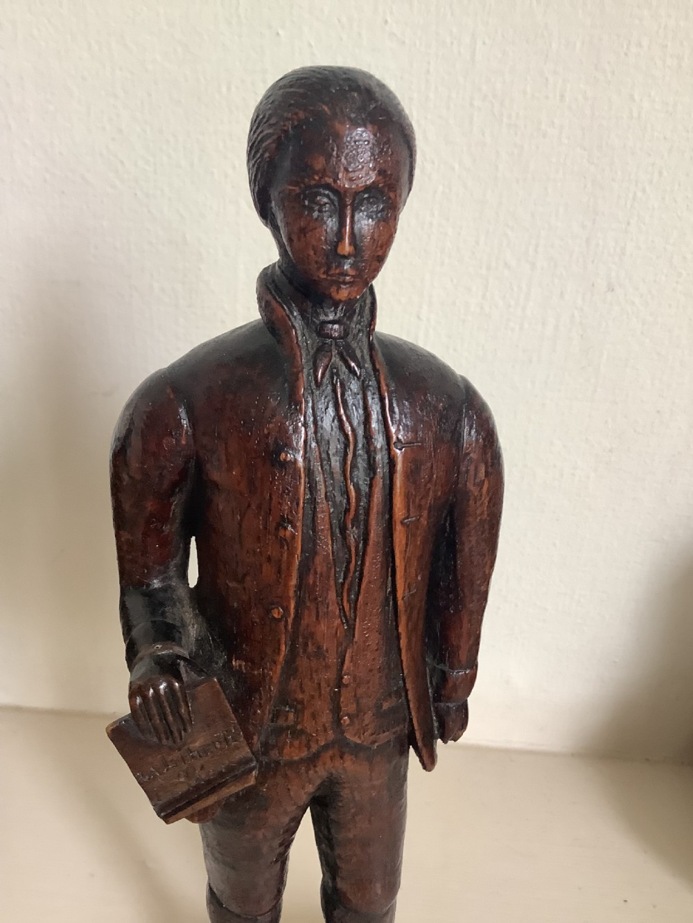 A dark hand-carved wood figure of an 18th-century man holding a book, standing on an integrated wooden base. View VI