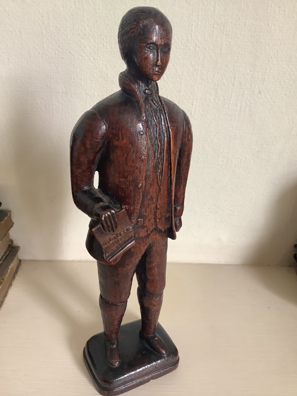A dark hand-carved wood figure of an 18th-century man holding a book, standing on an integrated wooden base. View VII