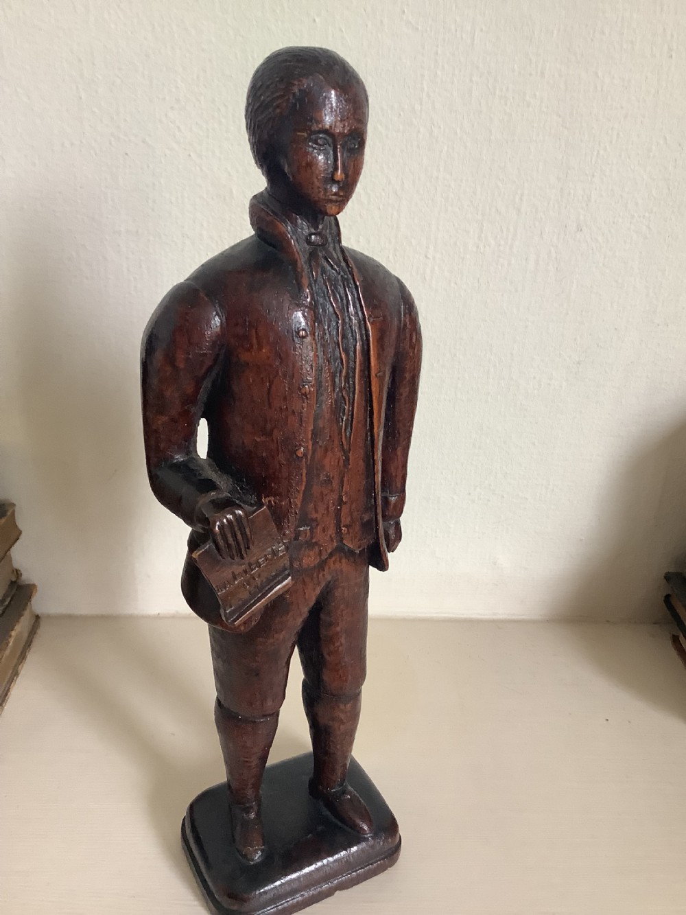 A dark hand-carved wood figure of an 18th-century man holding a book, standing on an integrated wooden base. View VIII