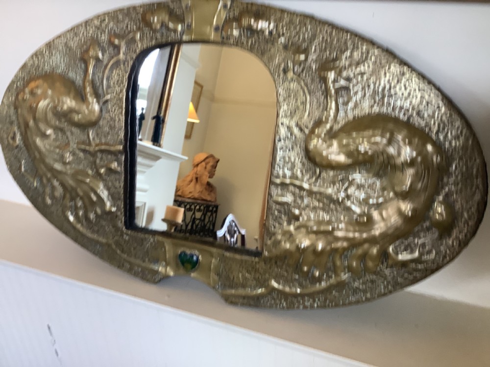 An oval Arts and Crafts hammered brass wall mirror featuring raised repousse peacocks flanking the central mirror plate and a green enamel cabochon at the bottom edge. View II