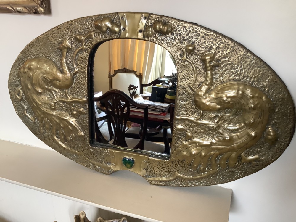 An oval Arts and Crafts hammered brass wall mirror featuring raised repousse peacocks flanking the central mirror plate and a green enamel cabochon at the bottom edge.