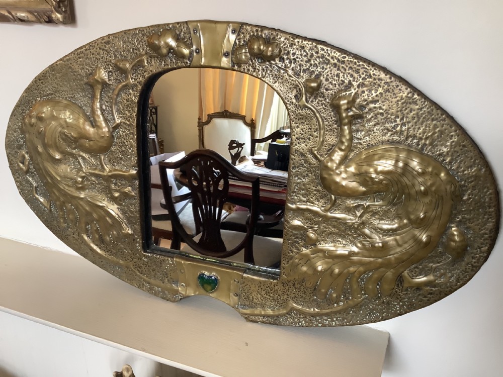 An oval Arts and Crafts hammered brass wall mirror featuring raised repousse peacocks flanking the central mirror plate and a green enamel cabochon at the bottom edge. View III