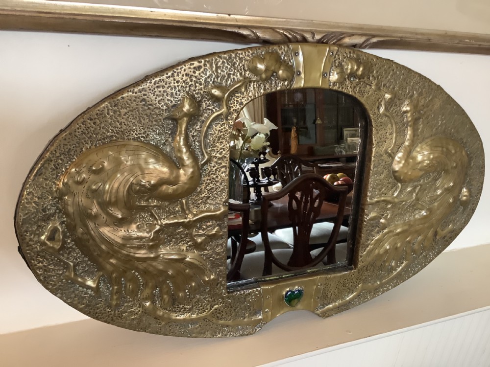 An oval Arts and Crafts hammered brass wall mirror featuring raised repousse peacocks flanking the central mirror plate and a green enamel cabochon at the bottom edge. View IV