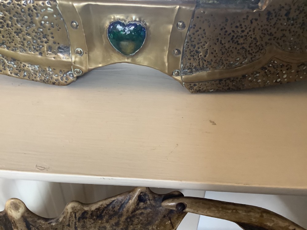 An oval Arts and Crafts hammered brass wall mirror featuring raised repousse peacocks flanking the central mirror plate and a green enamel cabochon at the bottom edge. View V