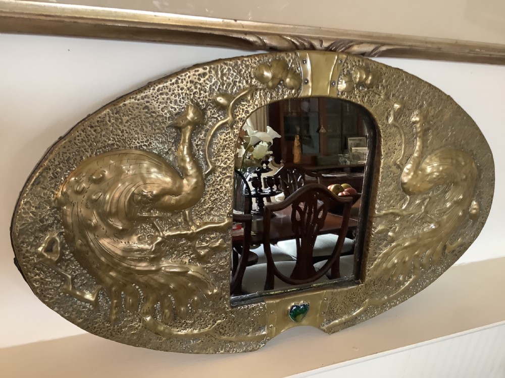 An oval Arts and Crafts hammered brass wall mirror featuring raised repousse peacocks flanking the central mirror plate and a green enamel cabochon at the bottom edge. View VII