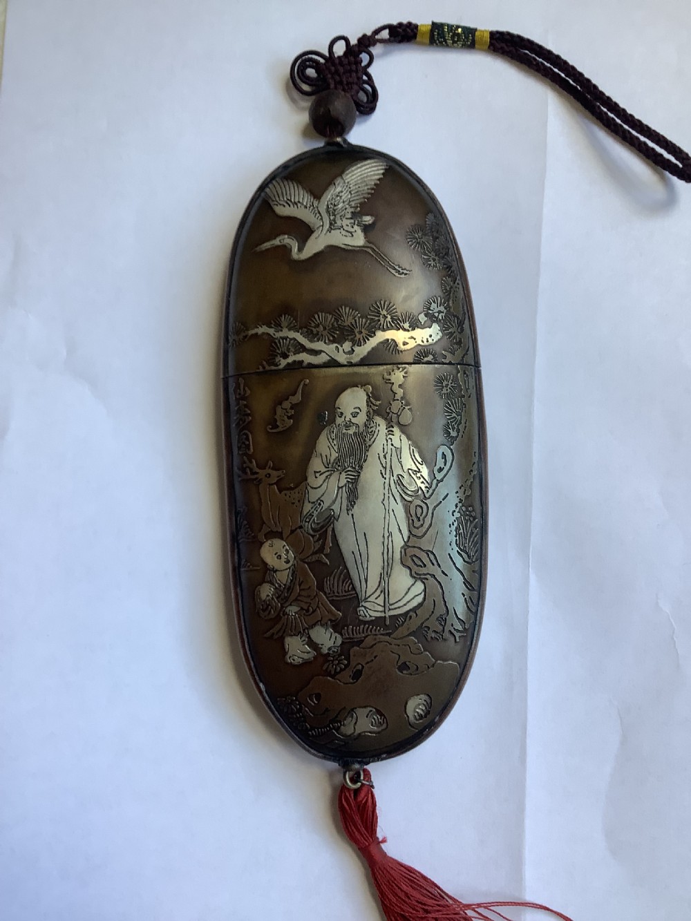 An oval dark bronze Japanese case featuring silver and mixed metal inlaid relief carvings of an elder, child, pine tree, and crane.