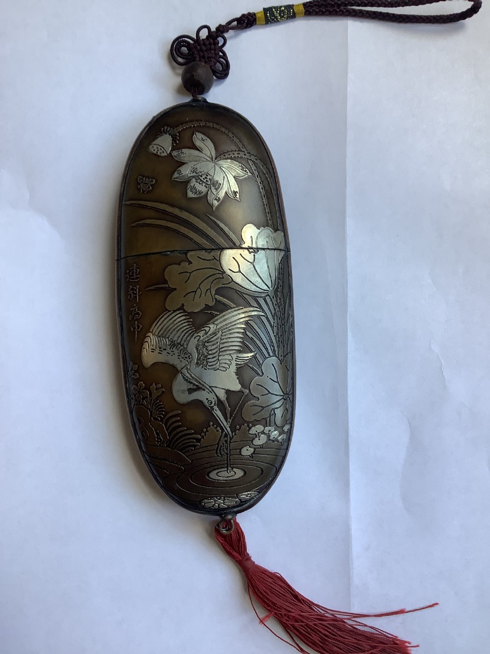 An oval dark bronze Japanese case featuring silver and mixed metal inlaid relief carvings of an elder, child, pine tree, and crane. View II