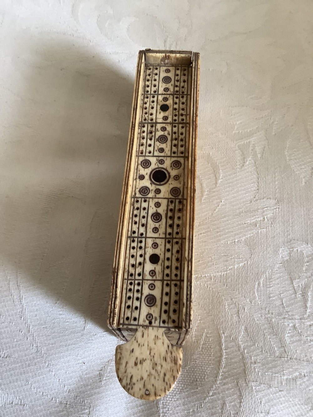 A 19th-century hand-carved bone domino set arranged on a white cloth alongside its decorated sliding wood box, cribbage scoring board, and carved pegs. View III