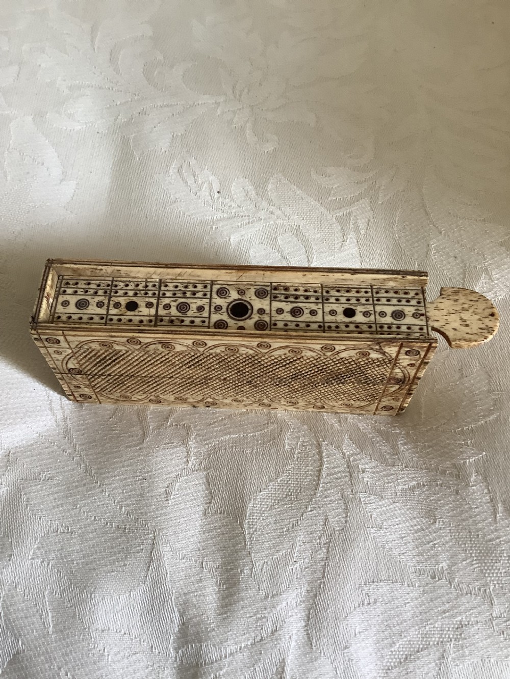 A 19th-century hand-carved bone domino set arranged on a white cloth alongside its decorated sliding wood box, cribbage scoring board, and carved pegs. View II