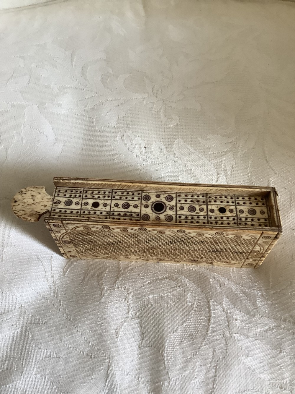 A 19th-century hand-carved bone domino set arranged on a white cloth alongside its decorated sliding wood box, cribbage scoring board, and carved pegs. View V