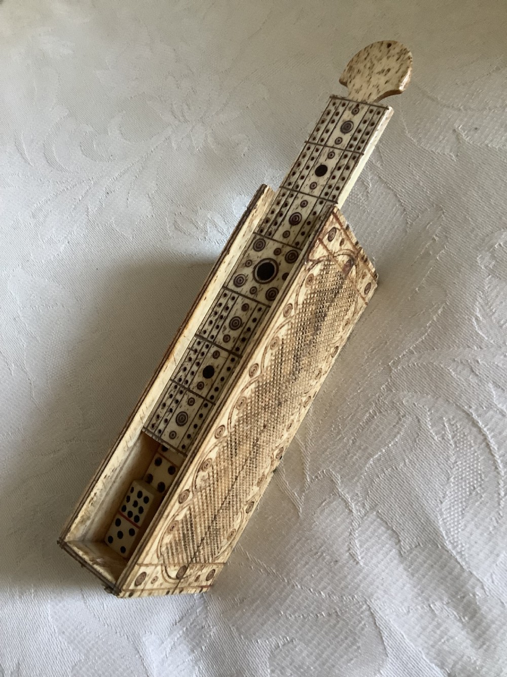 A 19th-century hand-carved bone domino set arranged on a white cloth alongside its decorated sliding wood box, cribbage scoring board, and carved pegs. View VI