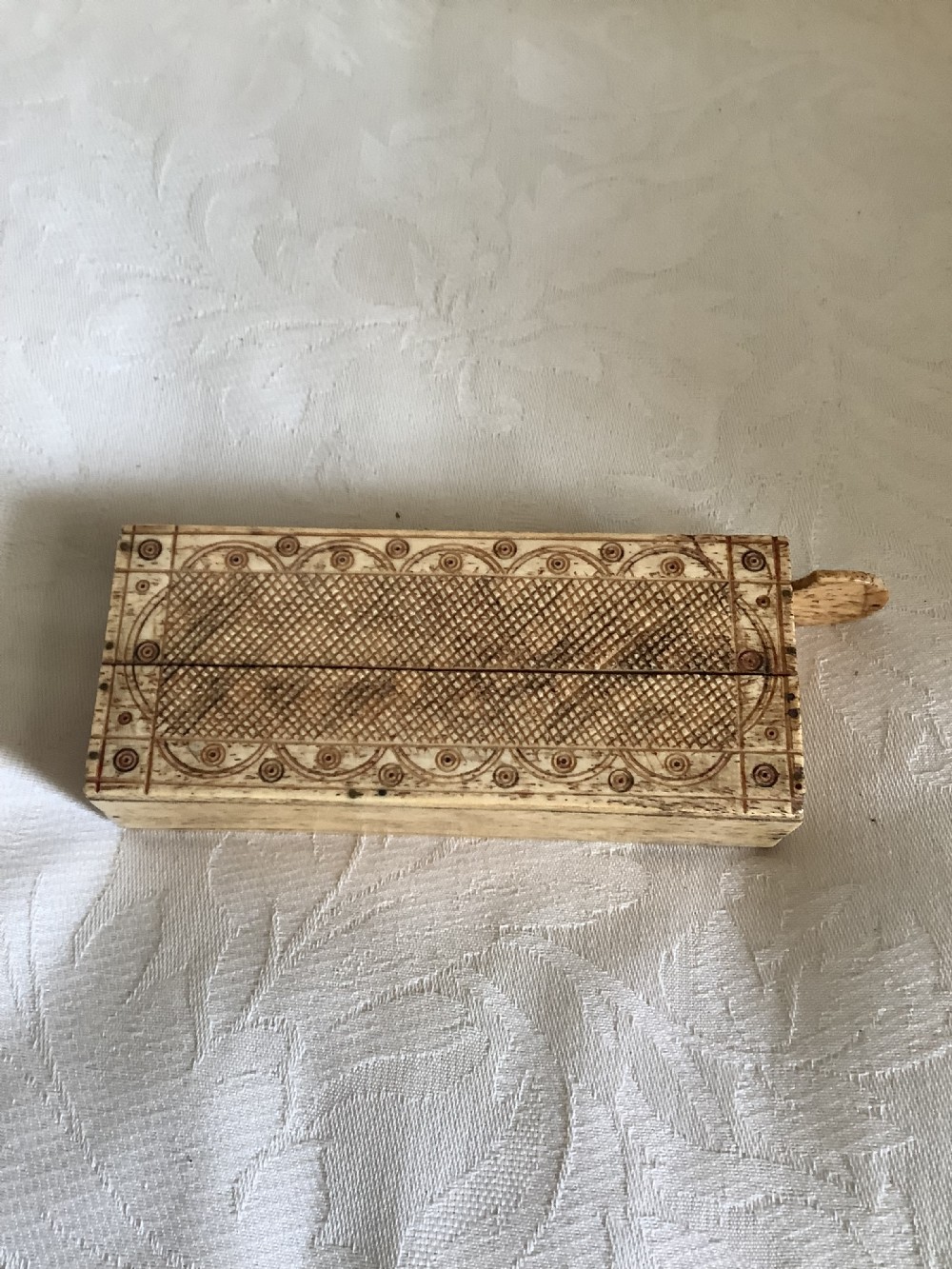 A 19th-century hand-carved bone domino set arranged on a white cloth alongside its decorated sliding wood box, cribbage scoring board, and carved pegs. View IX